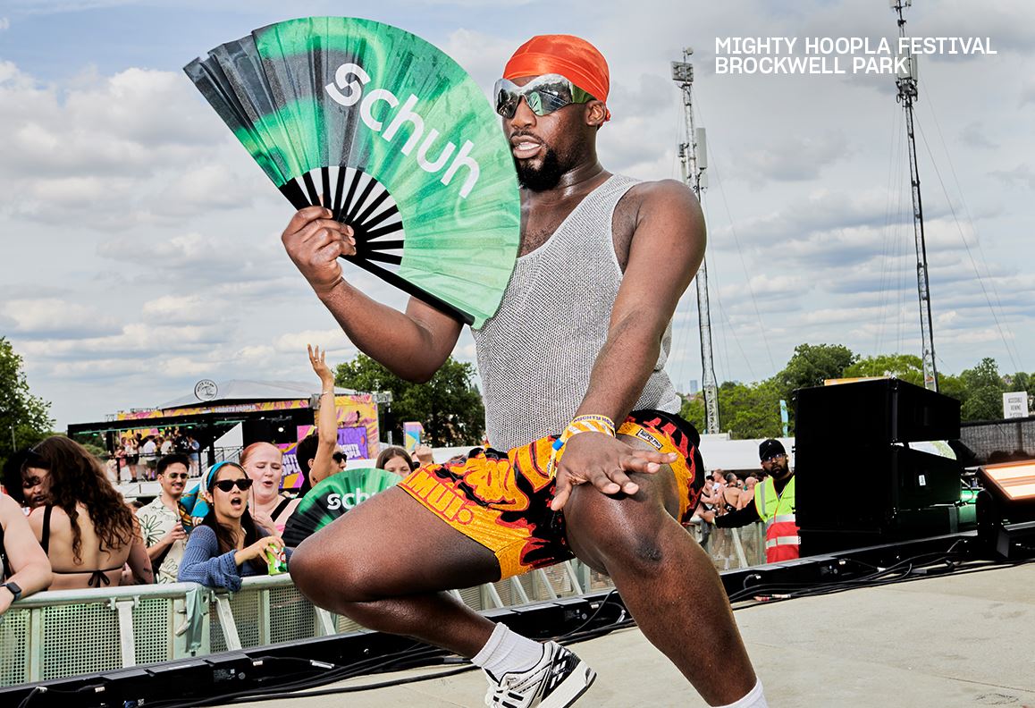 Image of a person posing with a green “schuh” fan at Mighty Hoopla Festival, wearing bold festival attire.